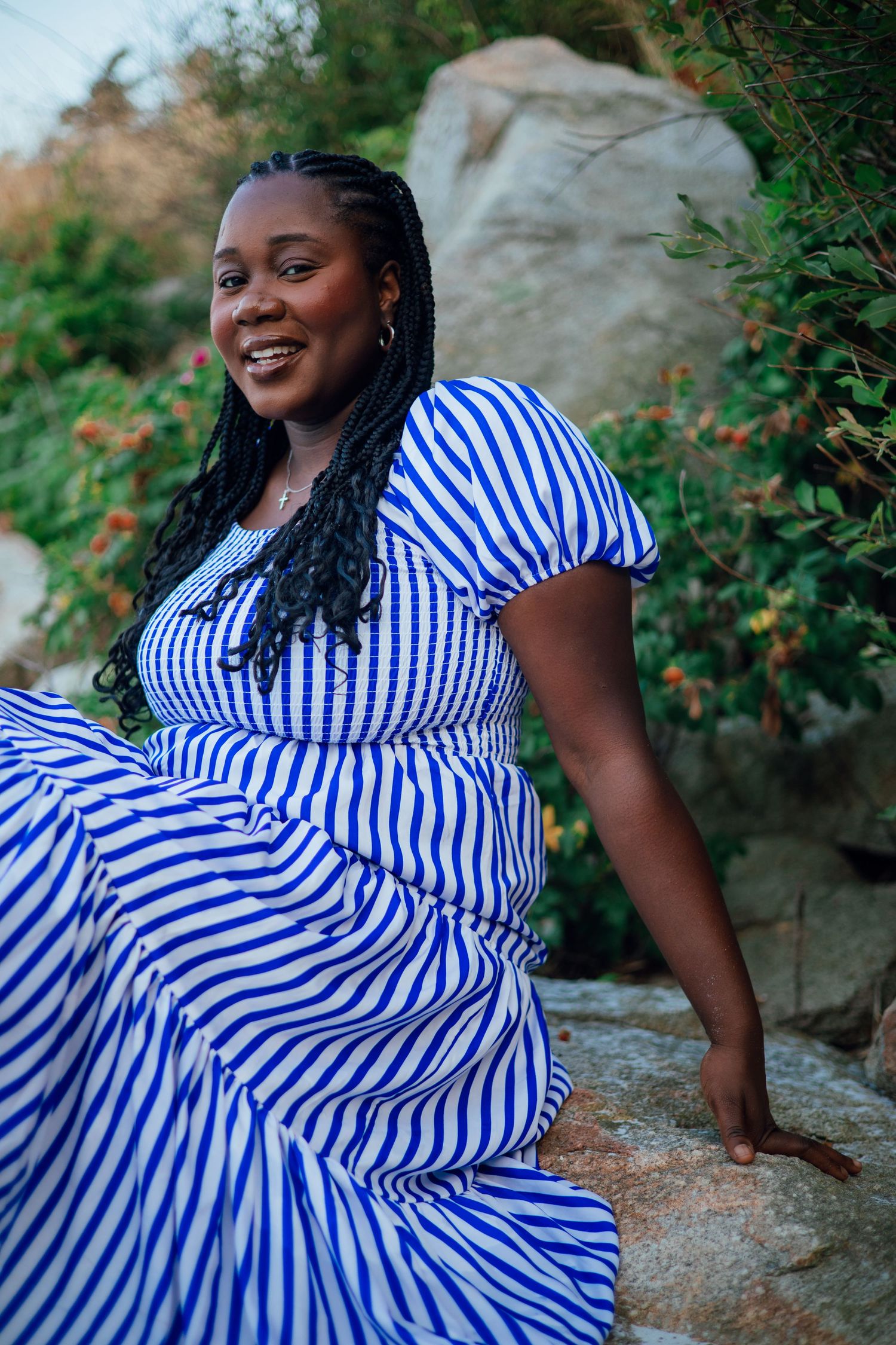 A smiling woman in a blue and white striped dress sits on rocks surrounded by lush greenery.