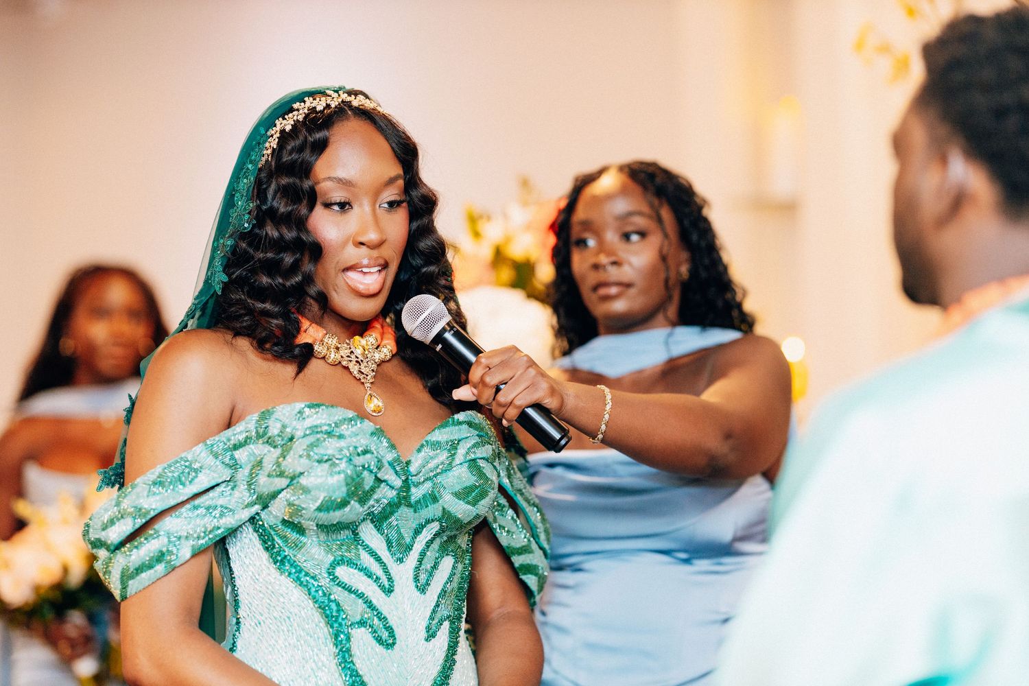 Bride in green embroidered gown reciting vows with microphone at wedding ceremony with bridesmaids in blue.
