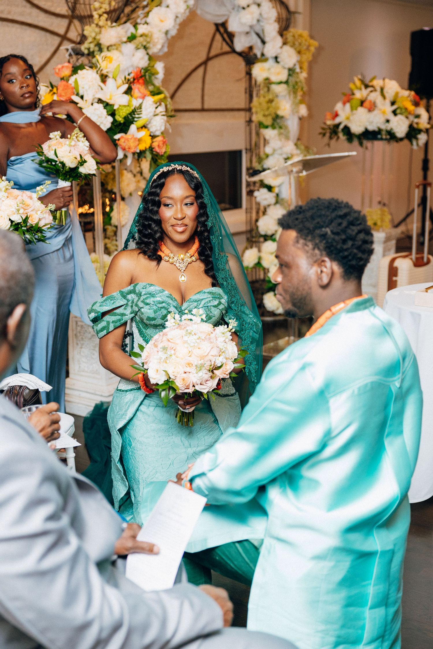 Bride in teal African wedding gown with veil holds bouquet during traditional ceremony.