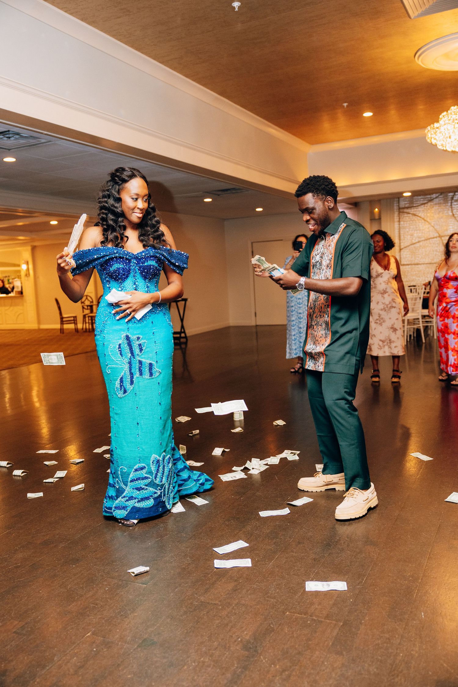 Bride in stunning turquoise beaded gown being sprayed with money during traditional African wedding reception dance.