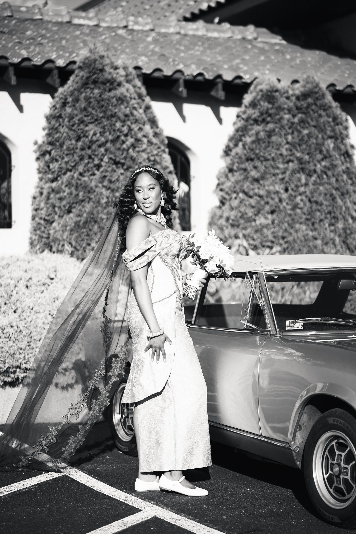 Black and white photo of bride in gown beside vintage car.