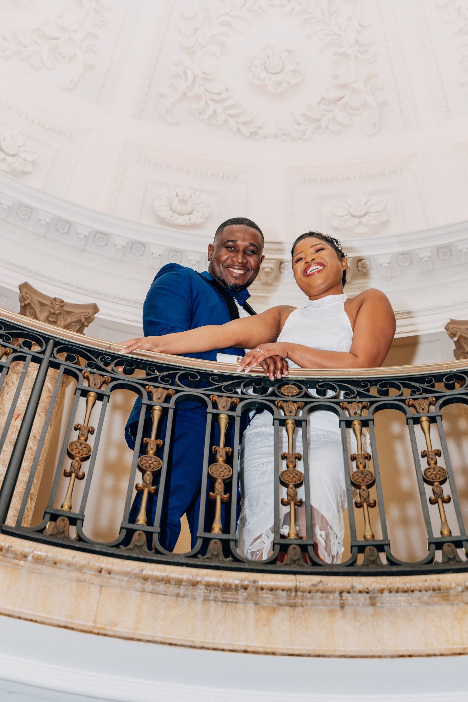 Newlyweds smiling on ornate balcony with decorative iron railing and domed ceiling.