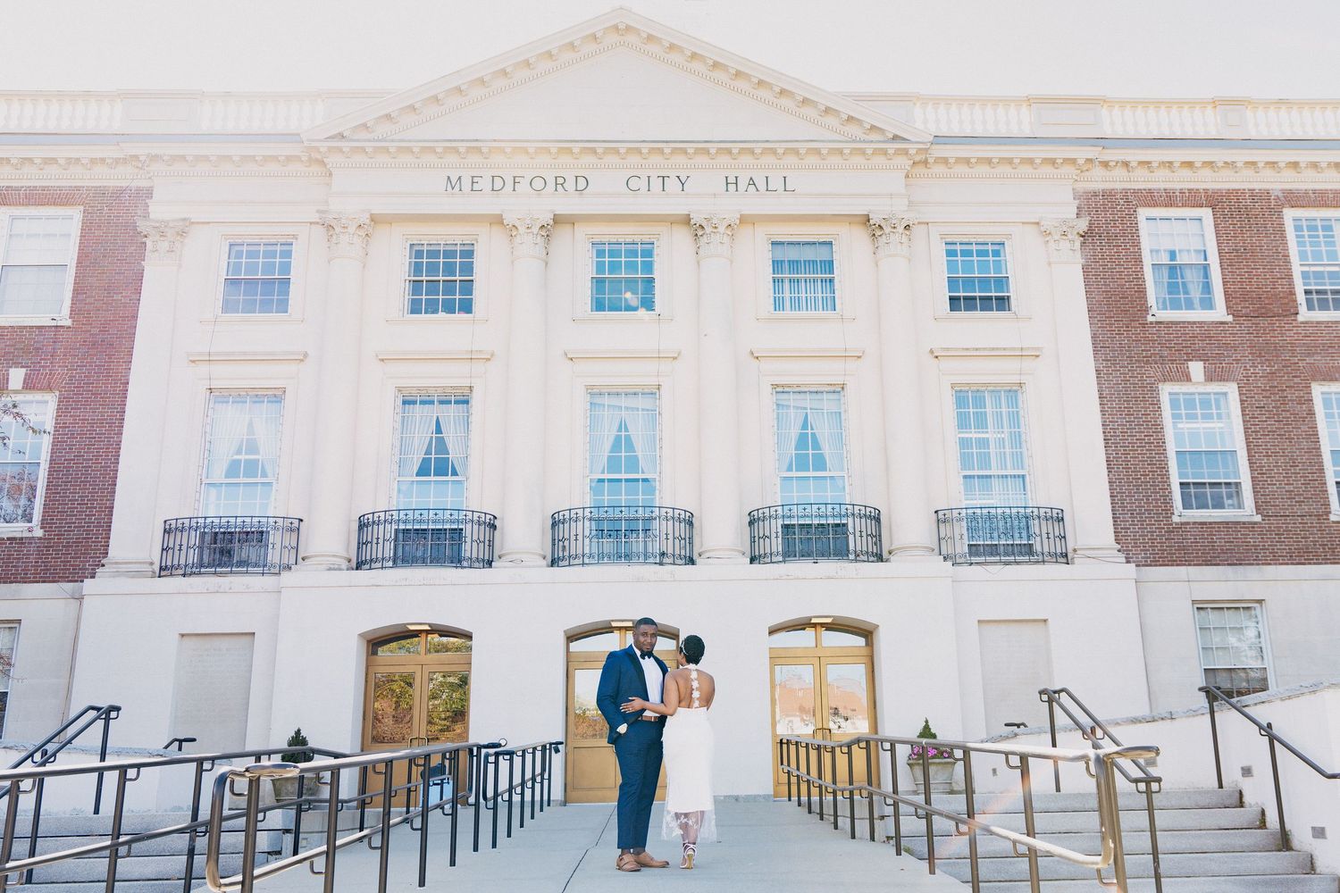 Couple shares a romantic kiss in front of Bedford City Hall, a grand brick building with white columns.