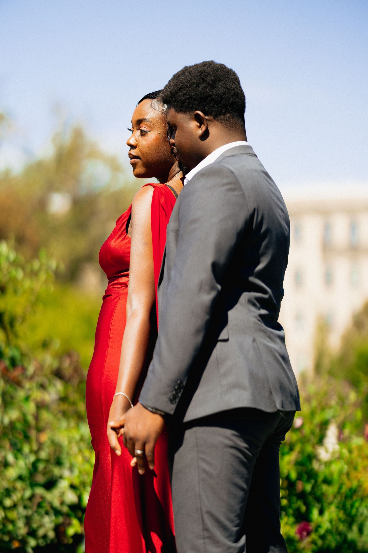 A couple stands close together in a garden, woman in red dress and man in gray suit holding hands.