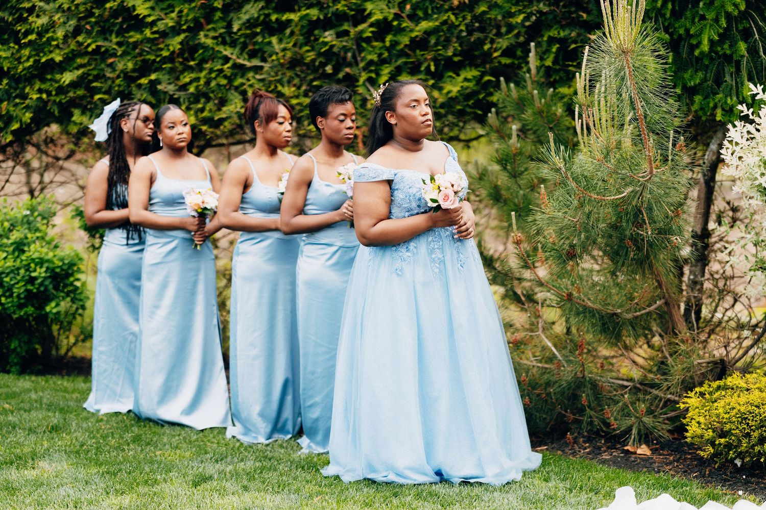 Four bridesmaids in matching light blue satin gowns holding bouquets standing at outdoor wedding ceremony.