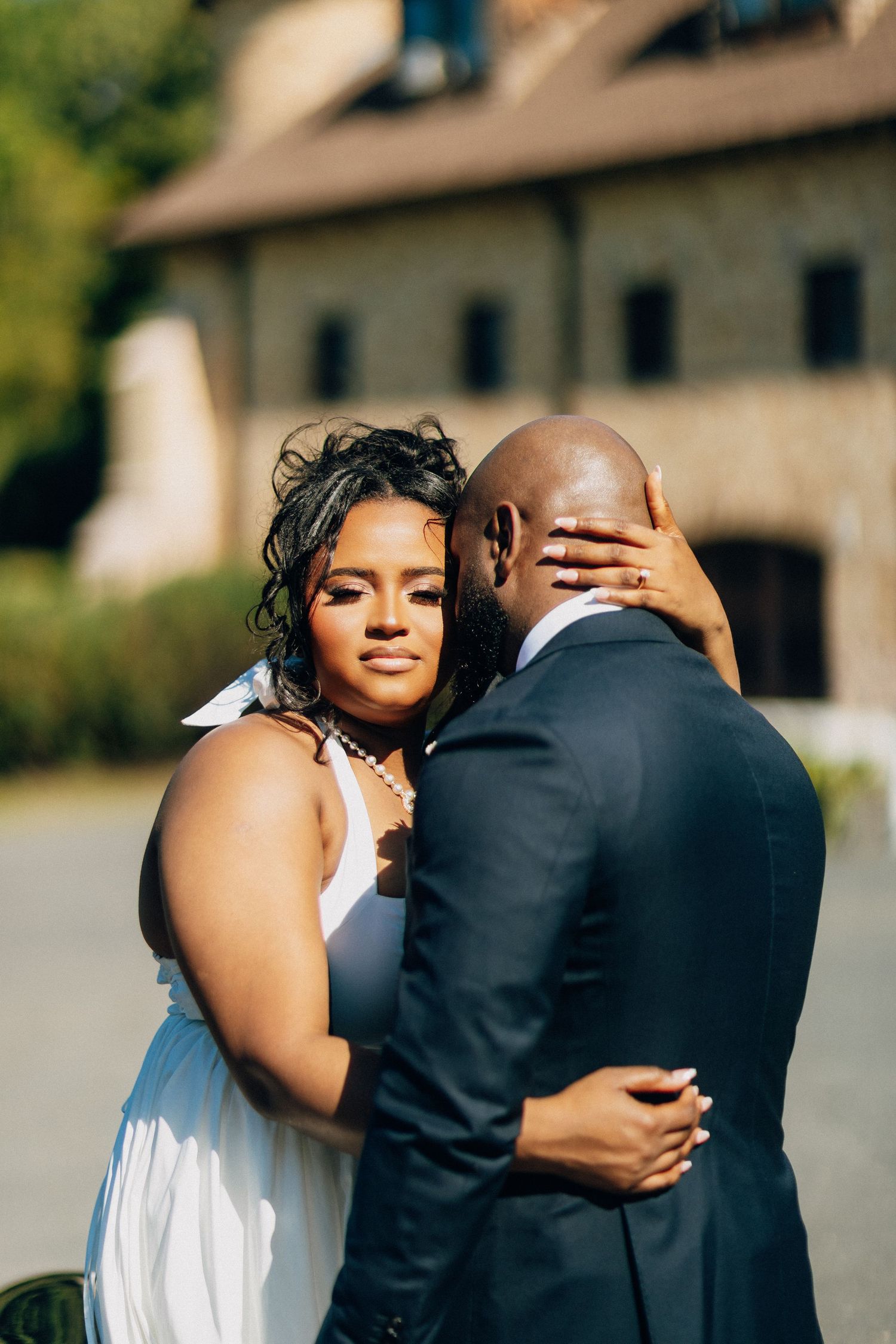 Beautiful bride in white gown embraces groom in navy suit outside rustic wedding venue.