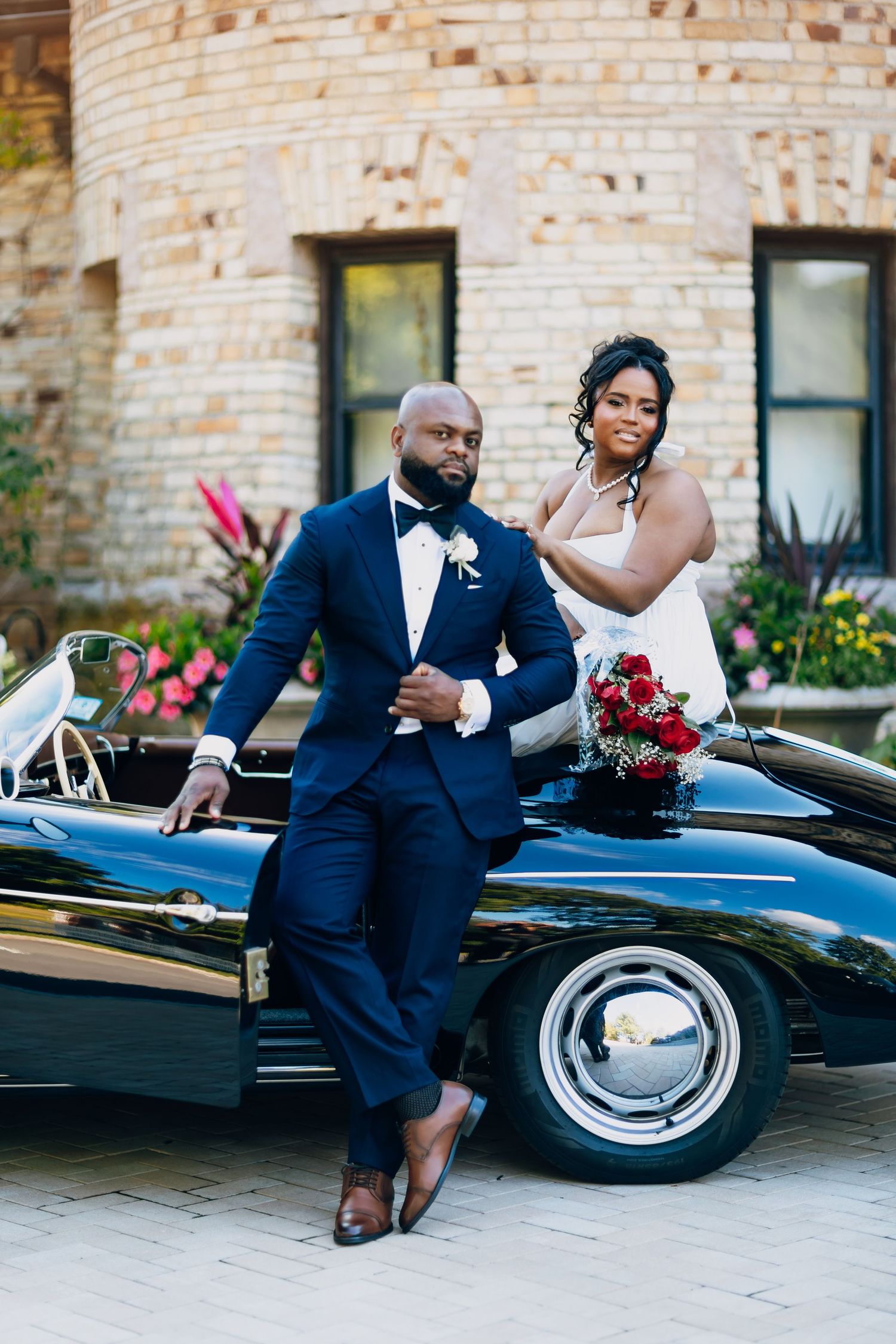 Bride and groom pose elegantly beside a classic black vintage convertible car on their wedding day.