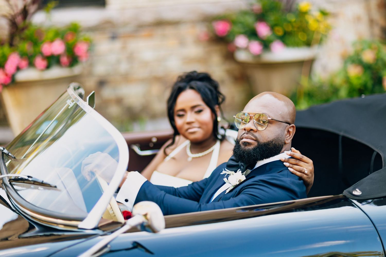 Newlyweds riding in a classic convertible car, bride embracing groom wearing gold sunglasses.