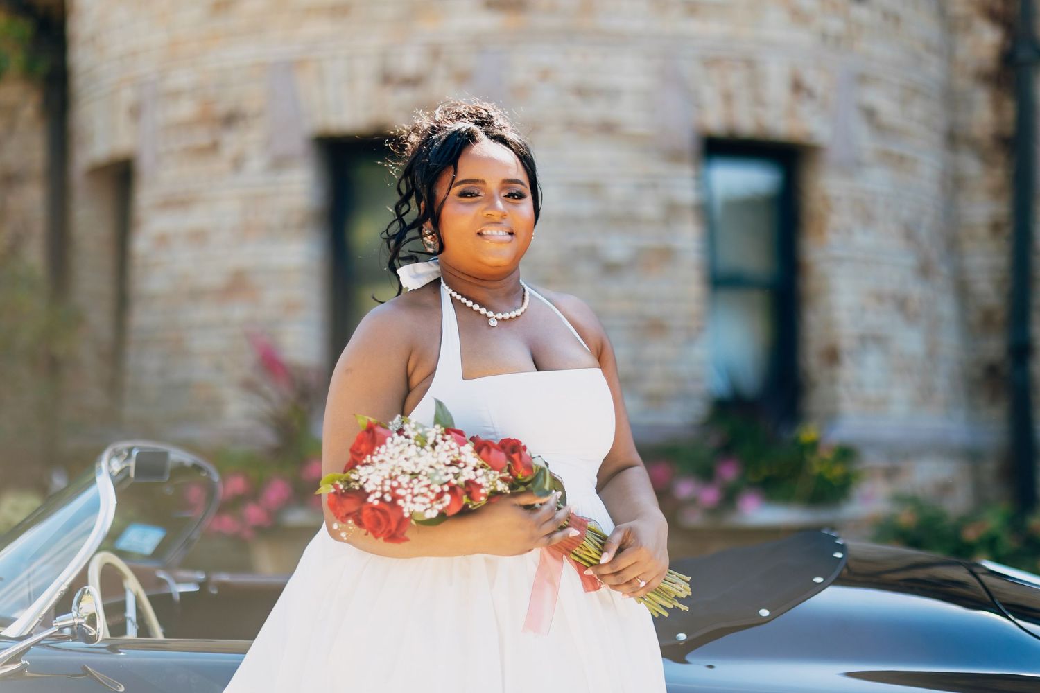 A radiant bride in a white halter dress holds a colorful bouquet beside a vintage car.
