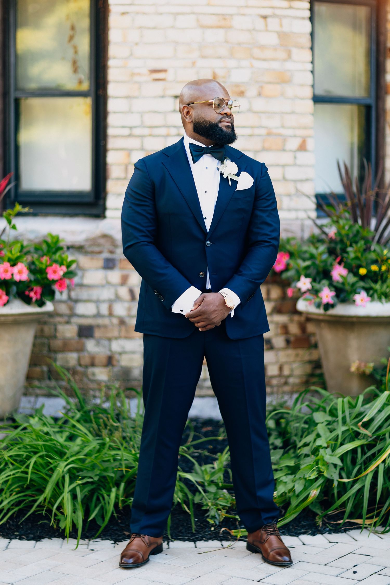 Handsome groom in navy tuxedo with white boutonniere stands before a brick building with flowers.