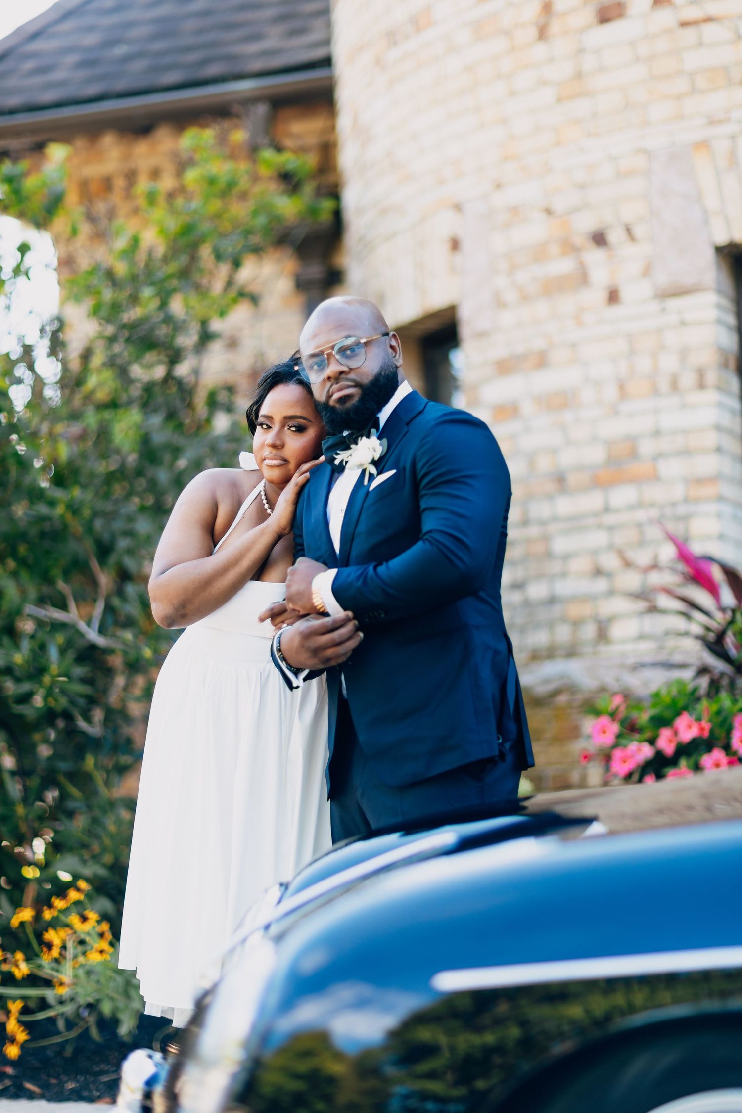 Bride and groom embrace near a vintage black car outside a stone building with colorful flowers.
