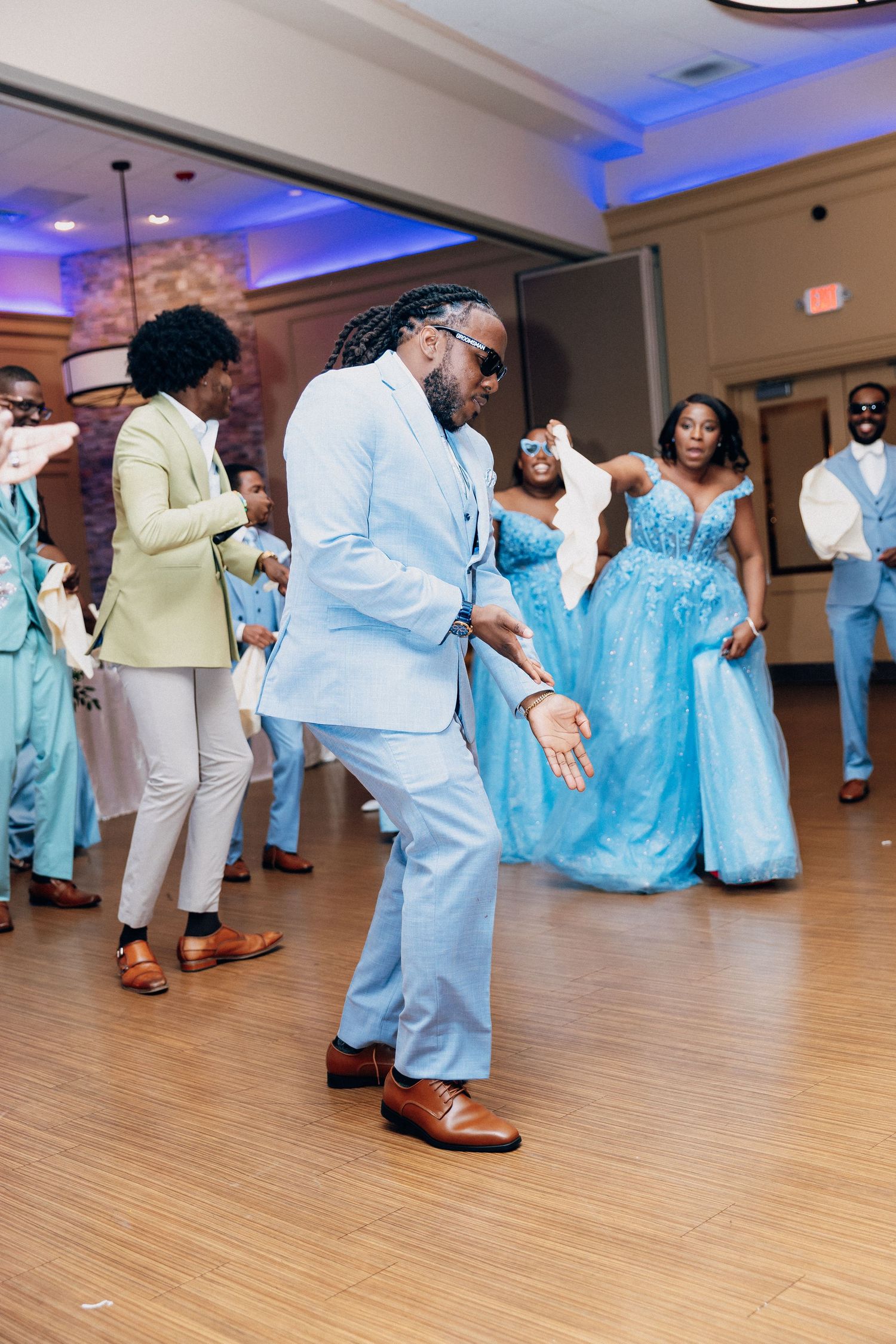 Guest in light blue suit and sunglasses dancing energetically at a lively wedding reception celebration.