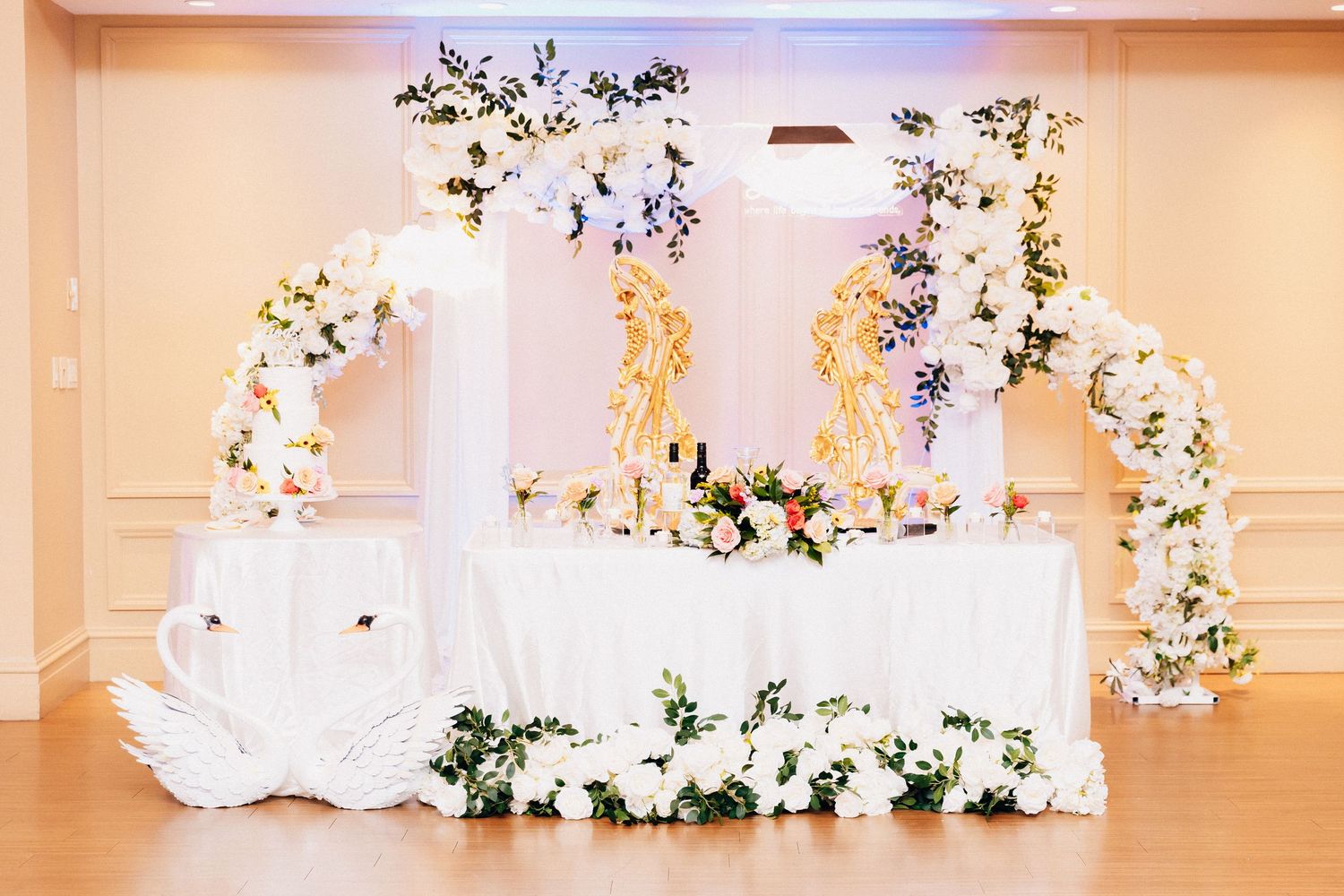 Elegant wedding sweetheart table with white floral arch, swan decorations, and lush flower arrangements in a ballroom.