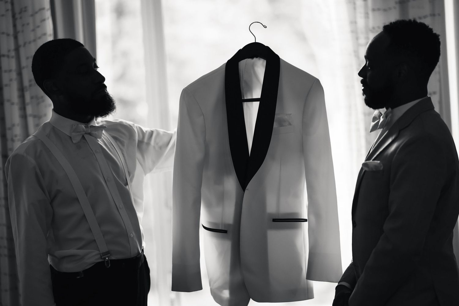 Two men in black and white photo examining a white tuxedo jacket on a hanger near a window.