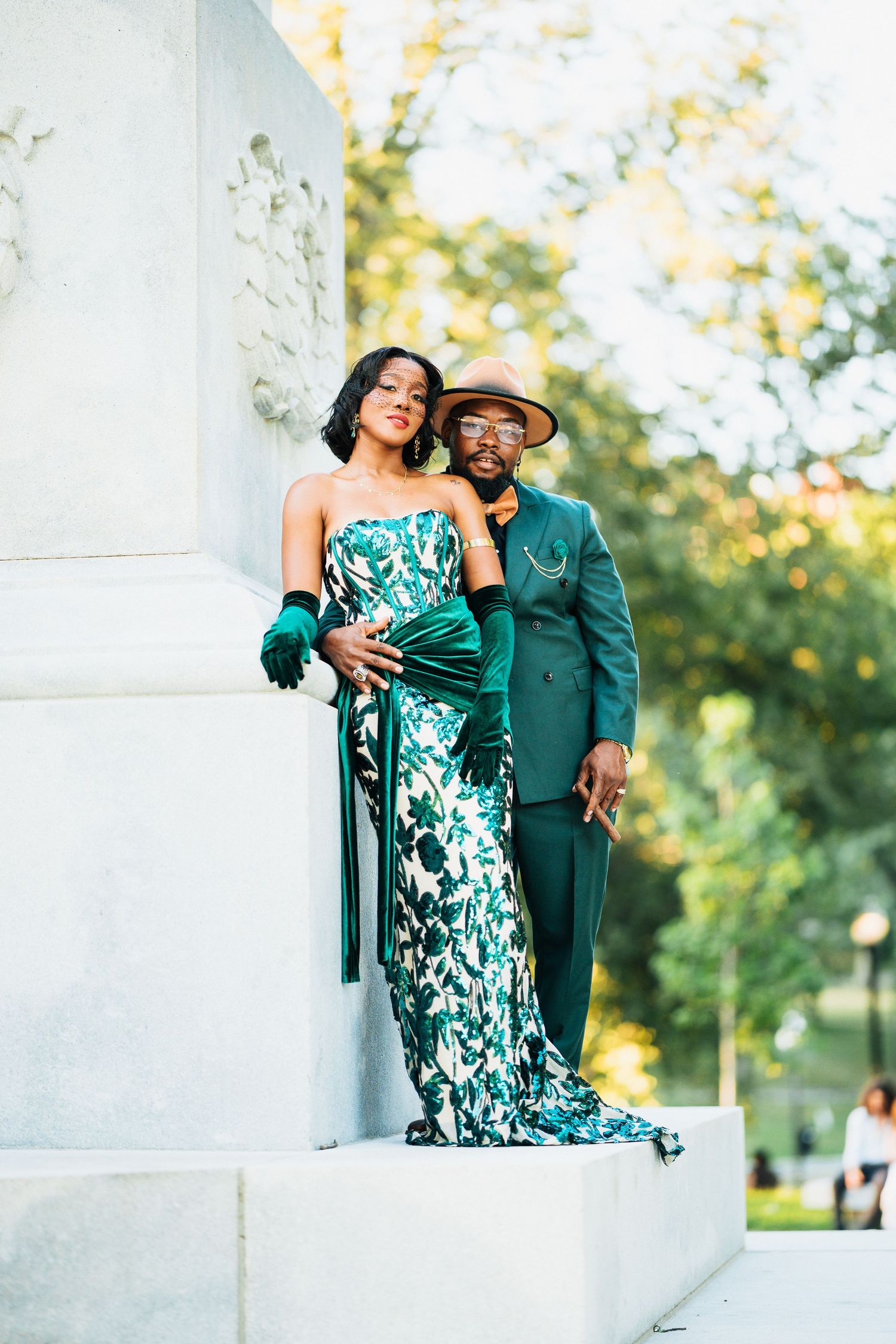 Couple in coordinating emerald green outfits posing against a white stone column.