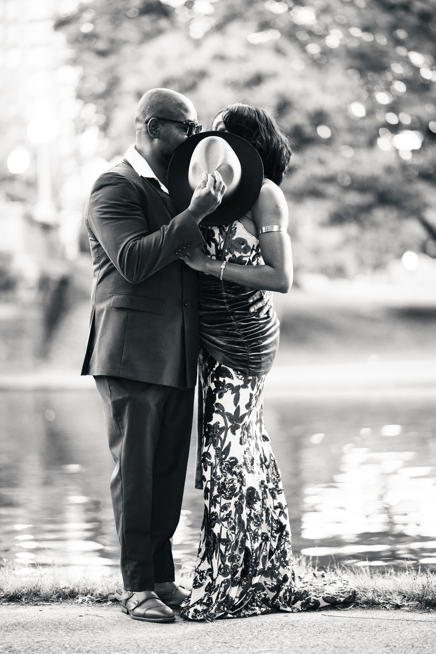 Black and white photo of couple kissing behind a wide-brim hat, wearing formal attire near a reflective pond.