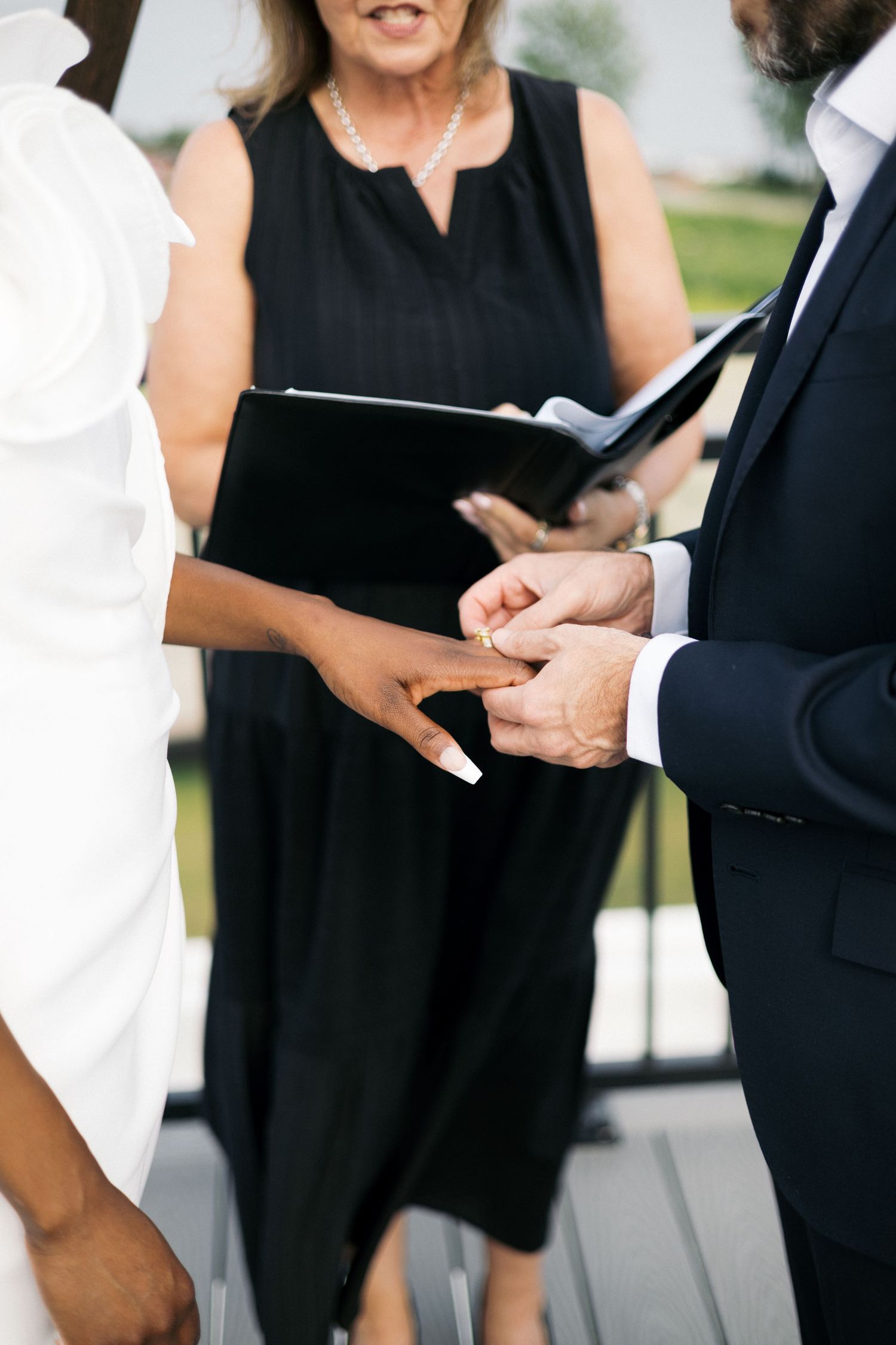 Groom placing a wedding ring on brides finger during an outdoor ceremony officiated by a woman in black.