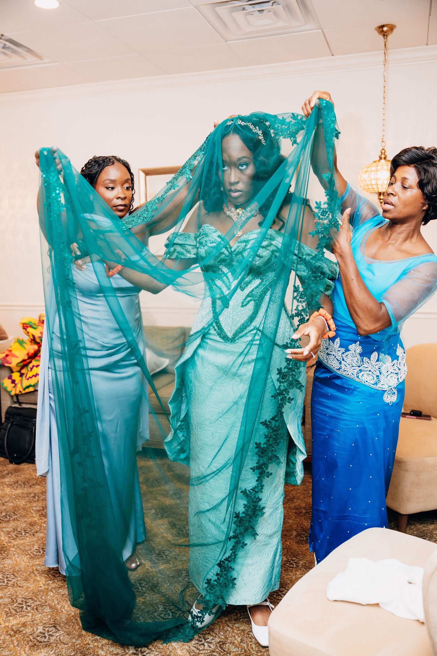 Two bridesmaids help a bride into a stunning teal lace wedding gown with an embroidered veil indoors.