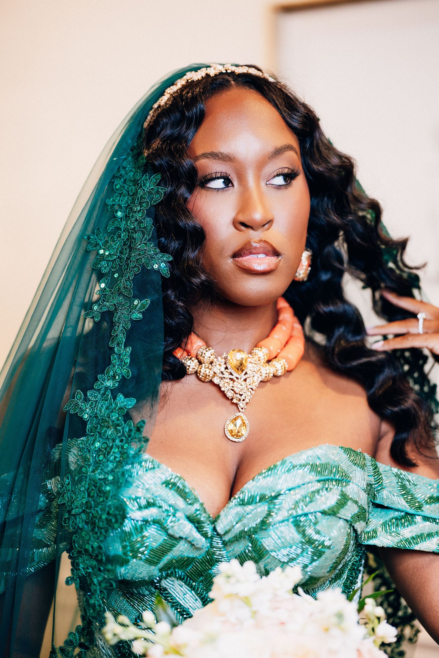 Close-up of African bride in teal off-shoulder gown with coral necklace, gold headpiece, and lace veil.