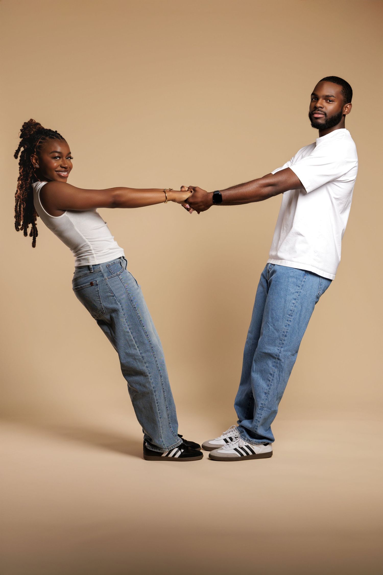 Young Black couple in matching white tees and jeans holding hands, leaning back playfully against beige background.