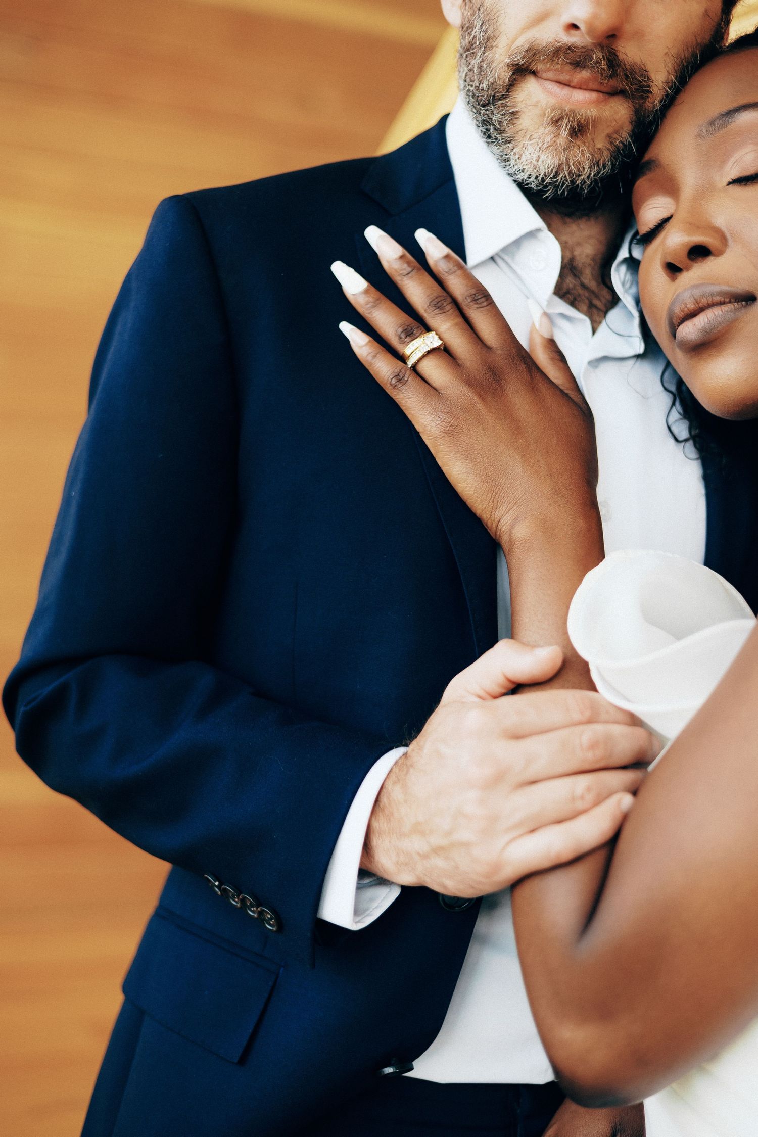 Interracial couple embracing on wedding day, bride in white dress with gold ring, groom in navy suit.