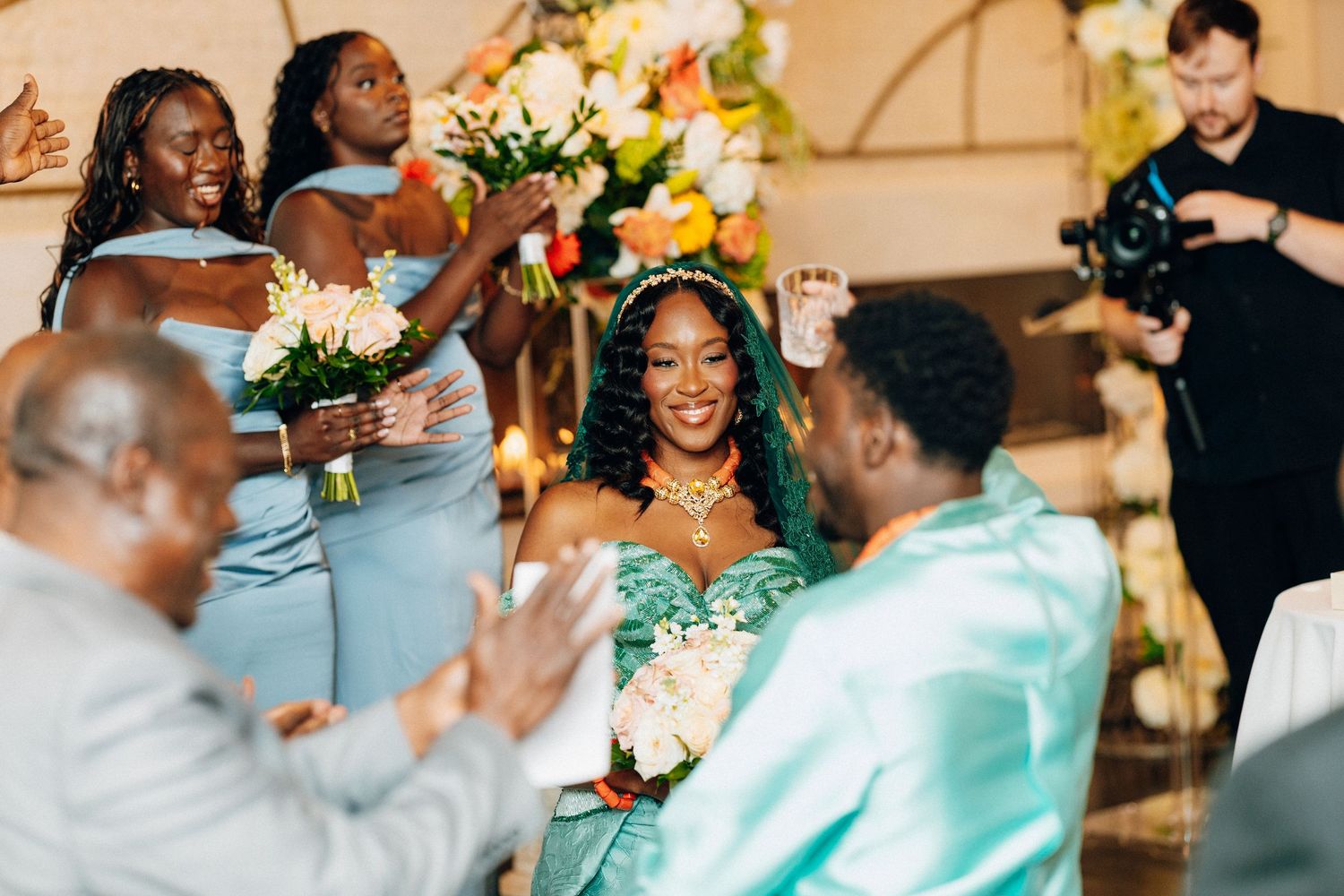 Bride in teal gown smiles at groom surrounded by bridesmaids in blue dresses at wedding.