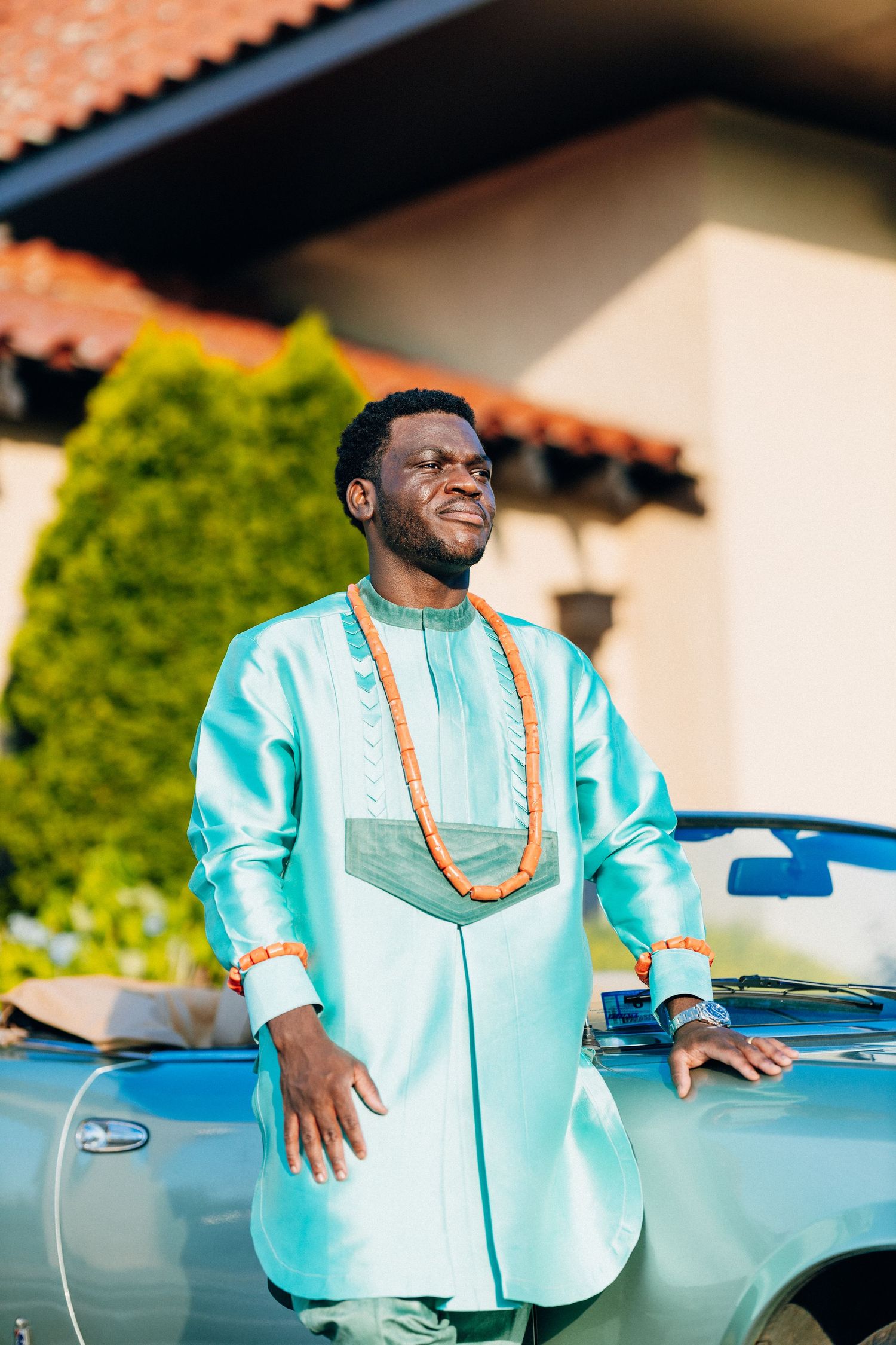 African groom in teal traditional attire smiles beside vintage blue car outdoors.