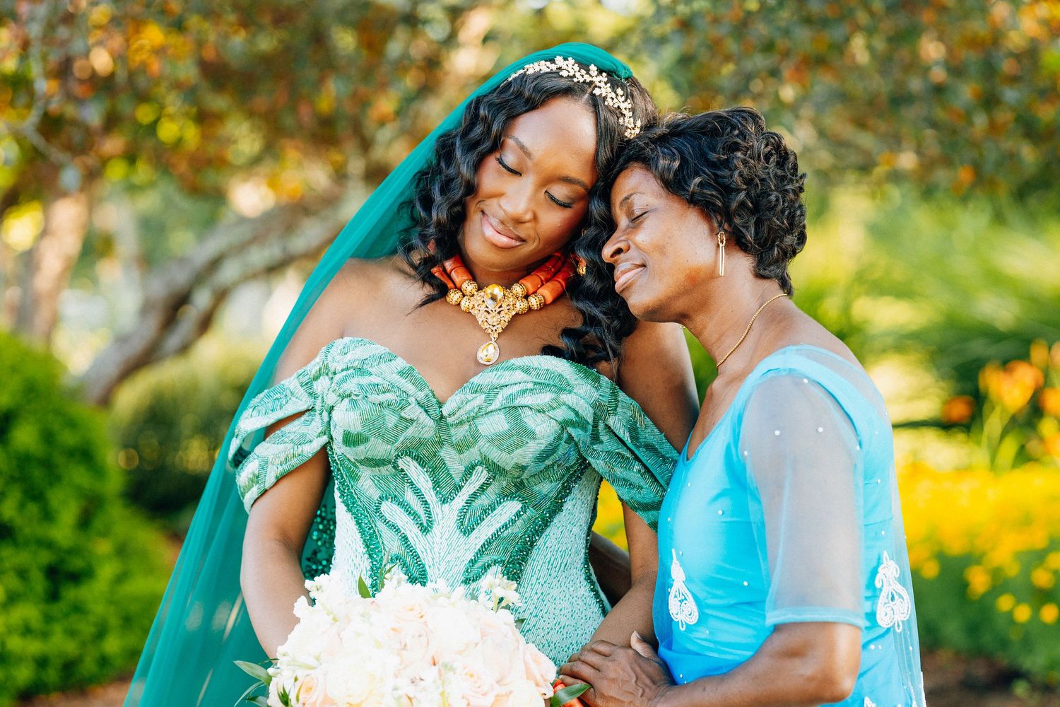 Bride in teal green lace wedding gown with veil and gold jewelry smiles with older woman in blue dress outdoors.