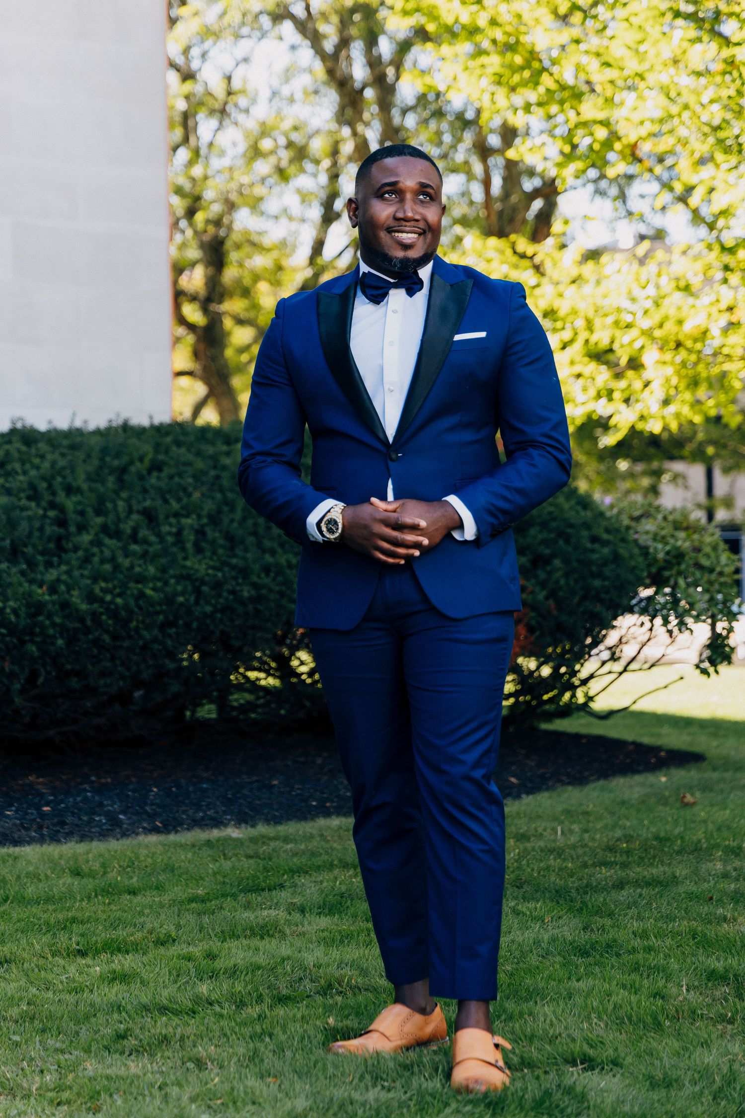 A well-dressed Black man in a navy blue tuxedo with bow tie and tan loafers smiling outdoors on green grass.