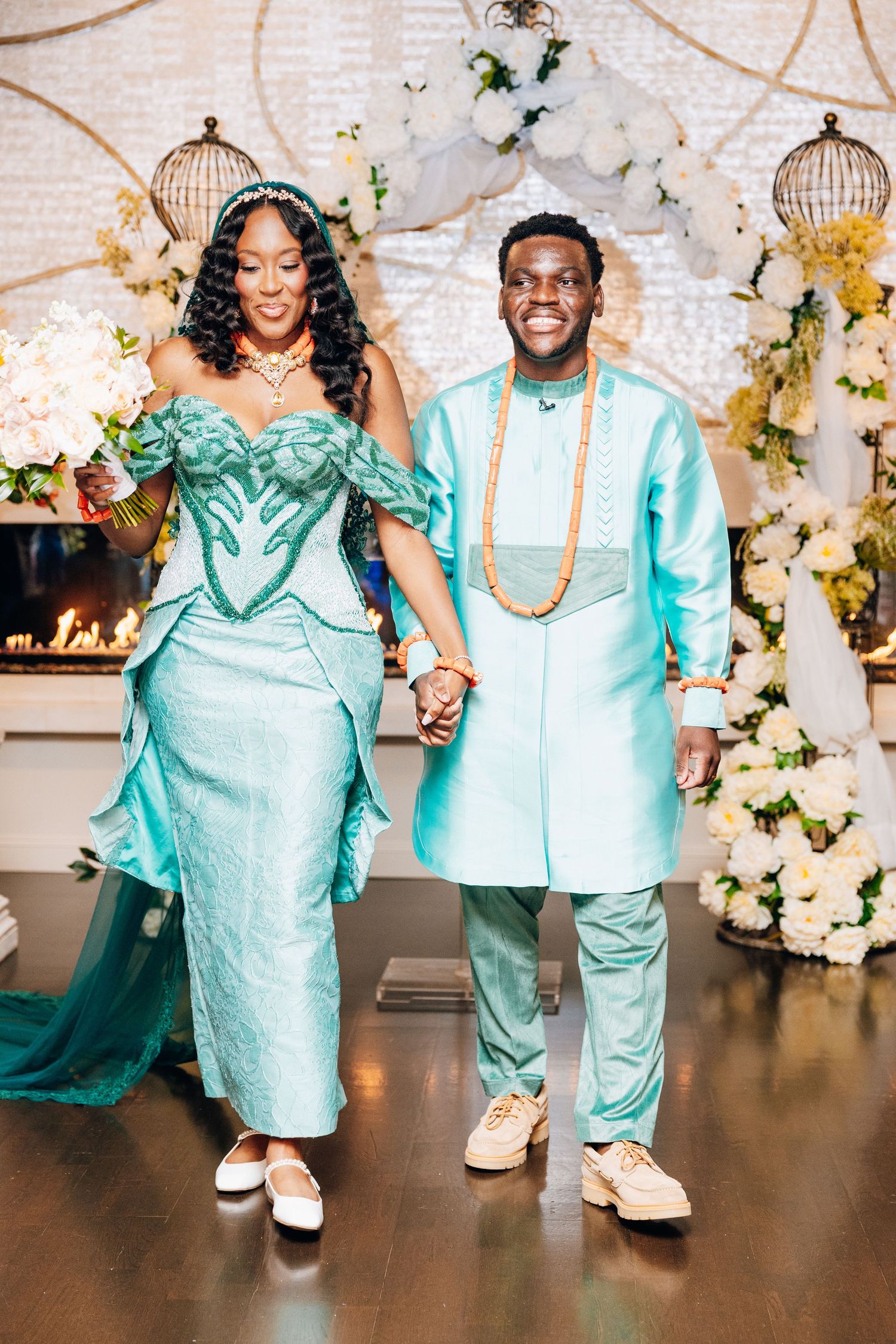 Nigerian couple in matching teal traditional wedding outfits hold hands before floral backdrop.