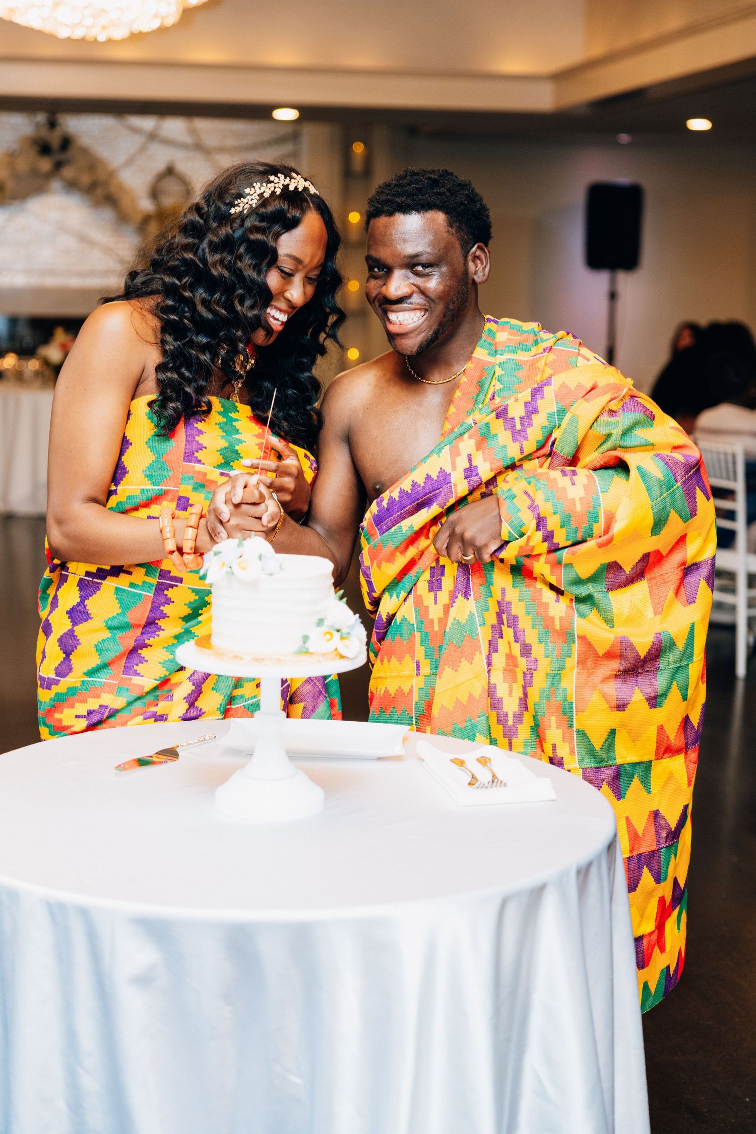 Couple in colorful Kente cloth attire cutting wedding cake at reception.