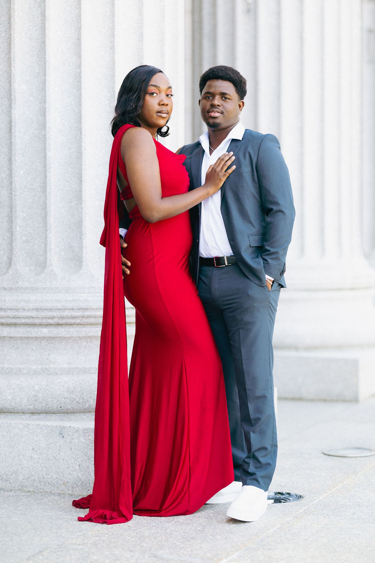 Elegant couple posing by marble columns, woman in red gown and man in gray suit.