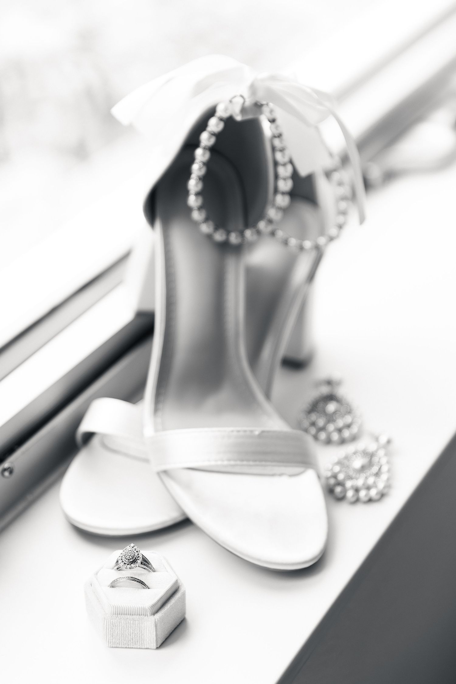 Black and white photo of bridal heels with pearl anklet, diamond ring in box, and pearl necklace on white surface.