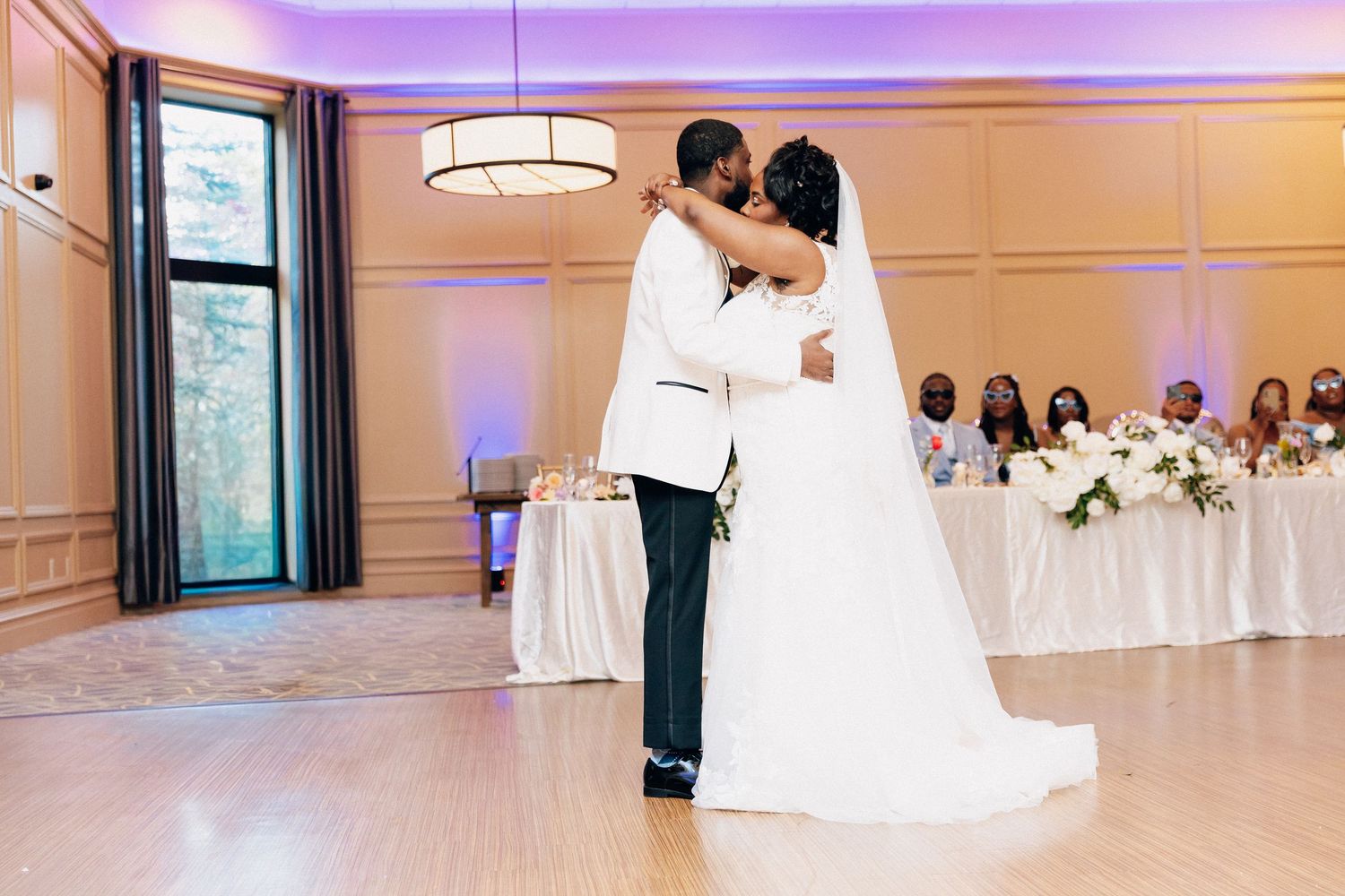 Bride and groom share their first dance in an elegant ballroom with purple uplighting and guests seated at decorated tables.