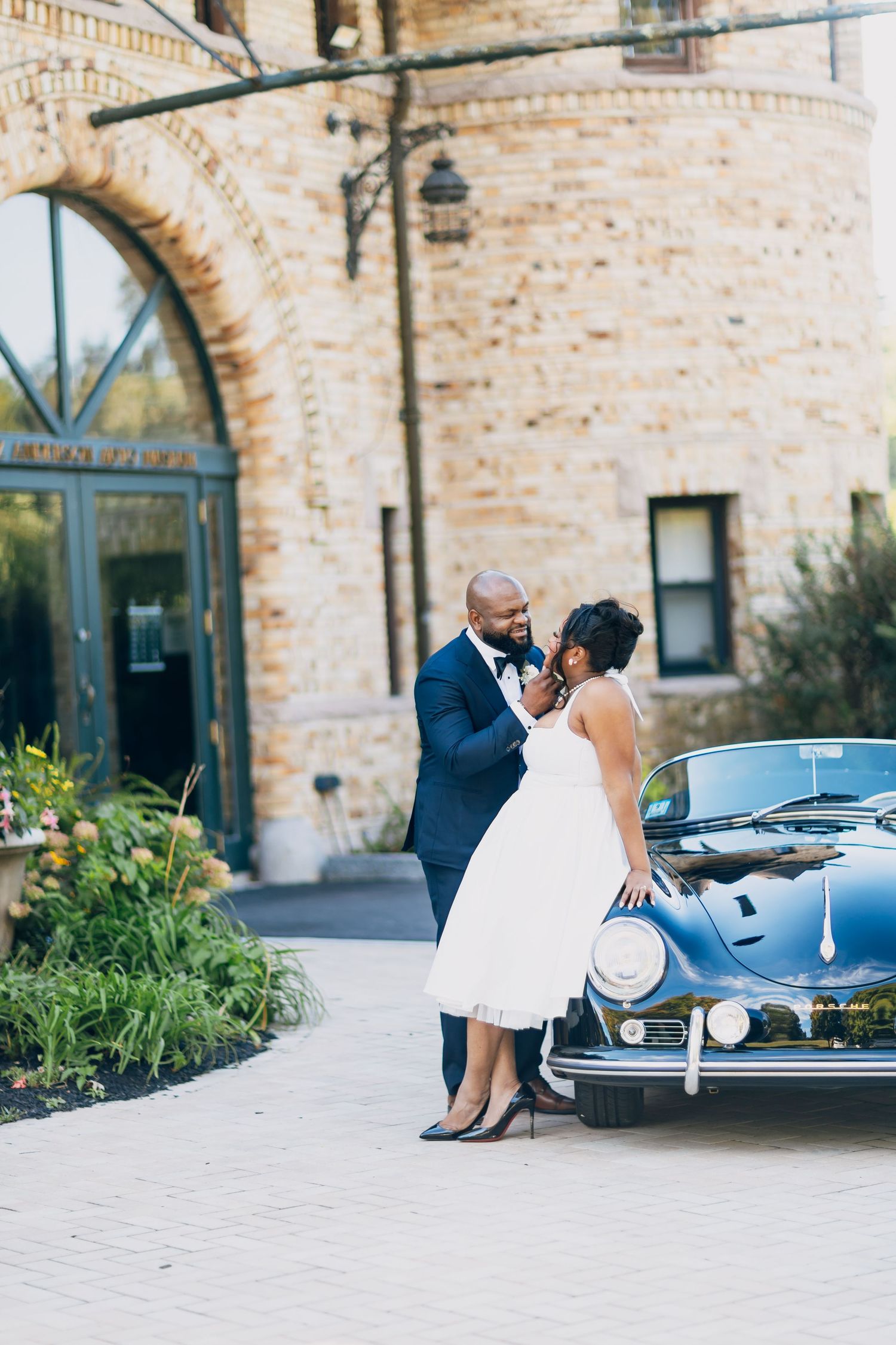 Bride and groom pose beside a vintage black car outside a brick building on their wedding day.
