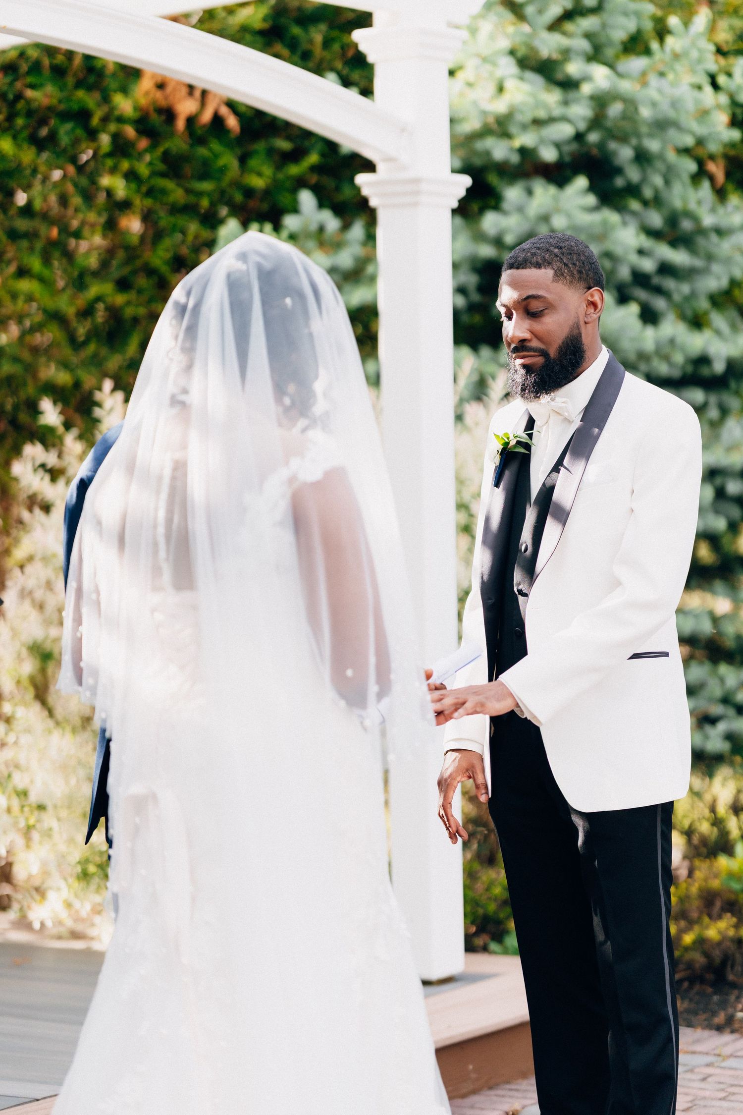 Groom in white tuxedo holds brides hand during outdoor wedding ceremony under white pergola.