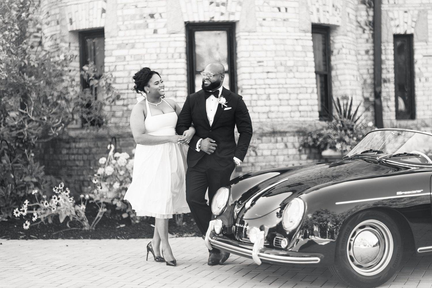 Bride and groom walking and laughing together beside a classic Porsche 356 at brick venue.