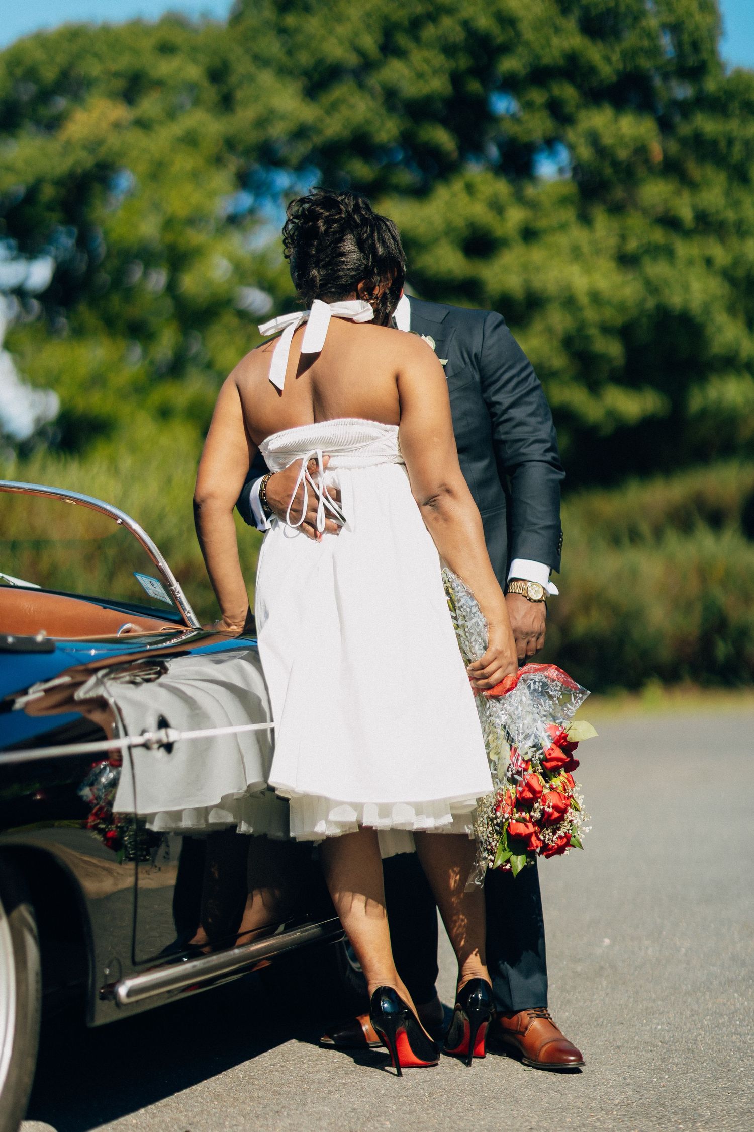 Bride in white halter dress and groom in navy suit stand by a vintage convertible car holding red bouquet.