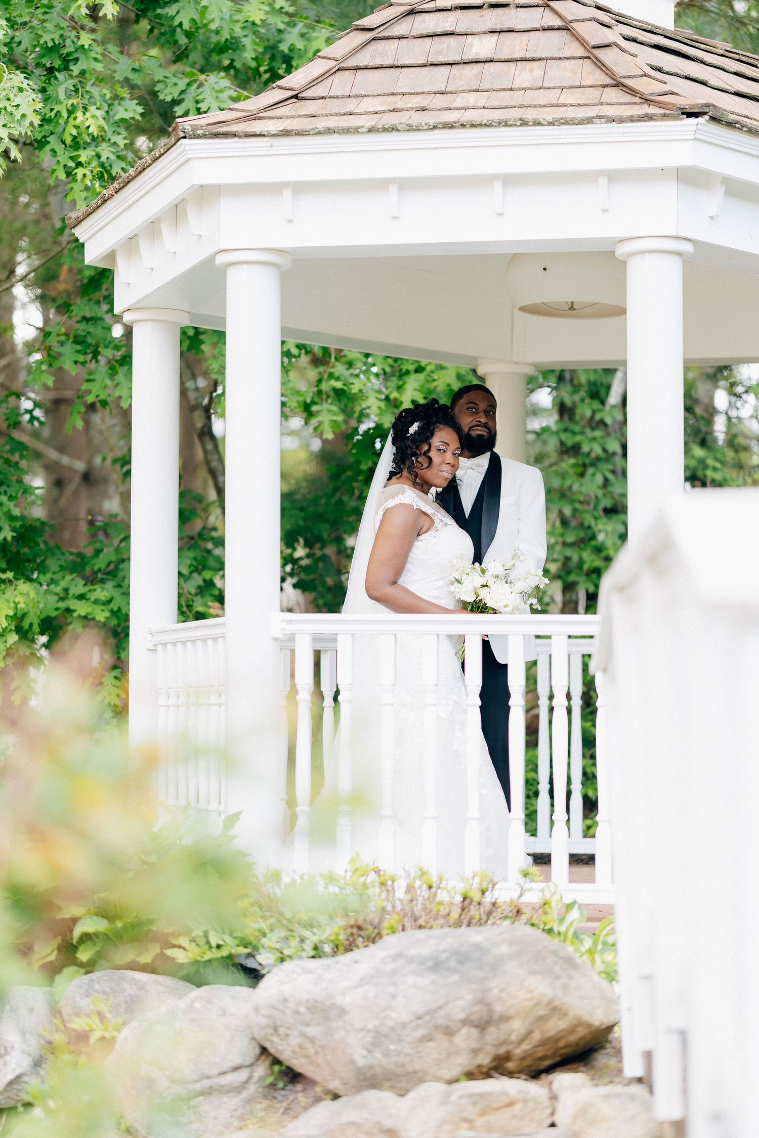 A bride and groom pose together in a white gazebo surrounded by lush greenery on their wedding day.