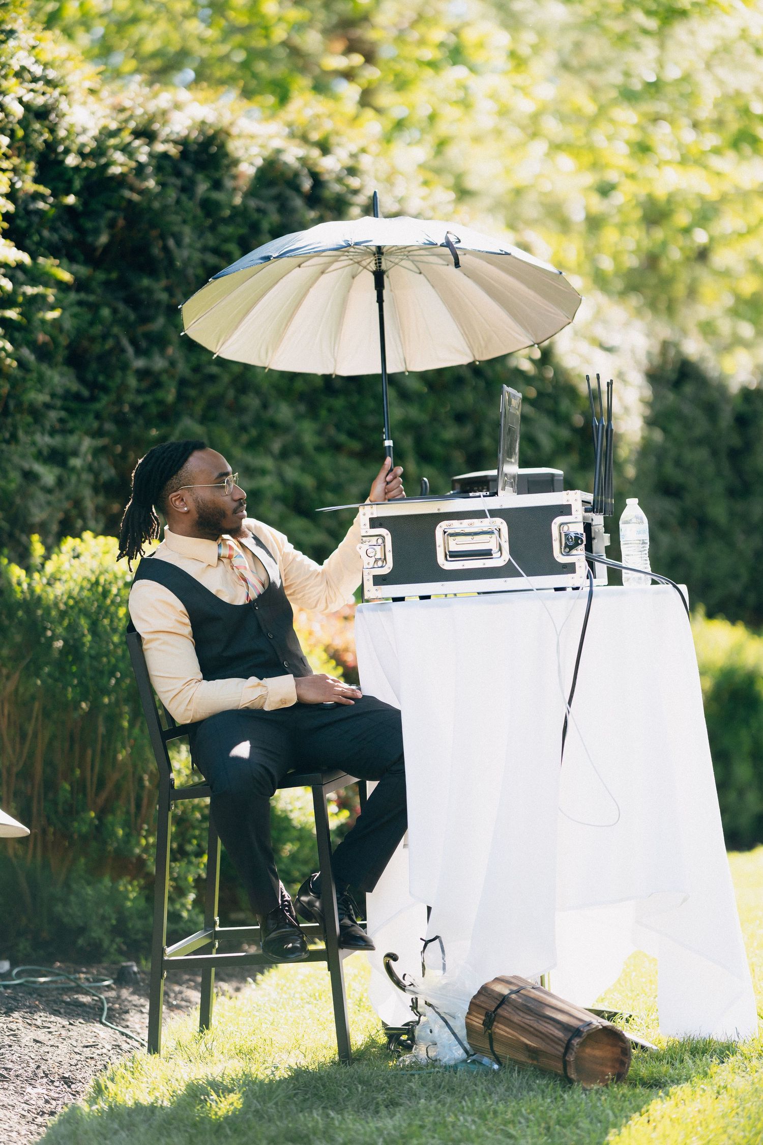 A stylish man with dreadlocks sits outdoors under a parasol umbrella next to a DJ setup at an outdoor event.