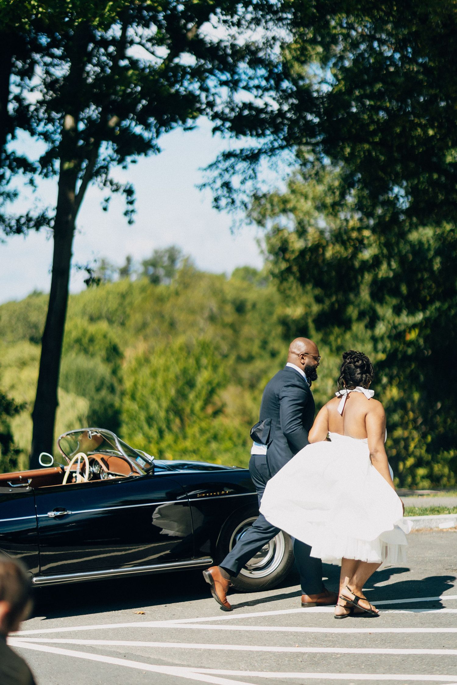 Bride in white midi dress and groom in navy suit walk toward a vintage black convertible on a sunny day.