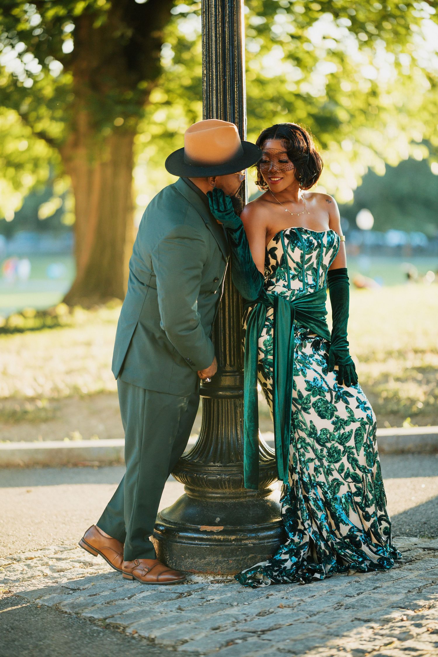 Stylish couple leaning against a lamppost in a park, man in green suit and hat, woman in floral dress.