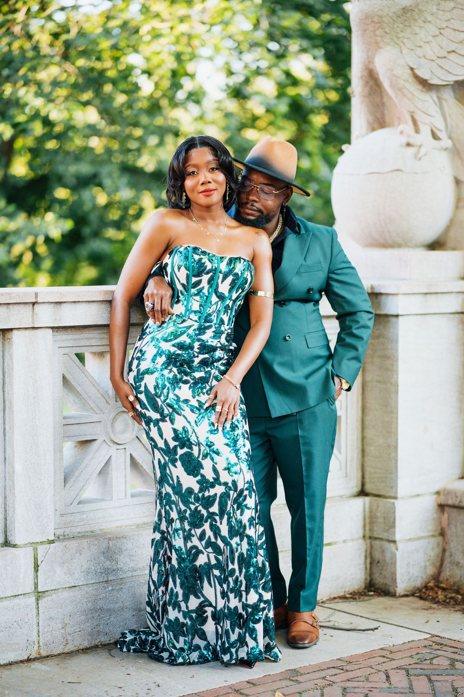 Elegant couple in matching green outfits posing by stone balustrade in park.