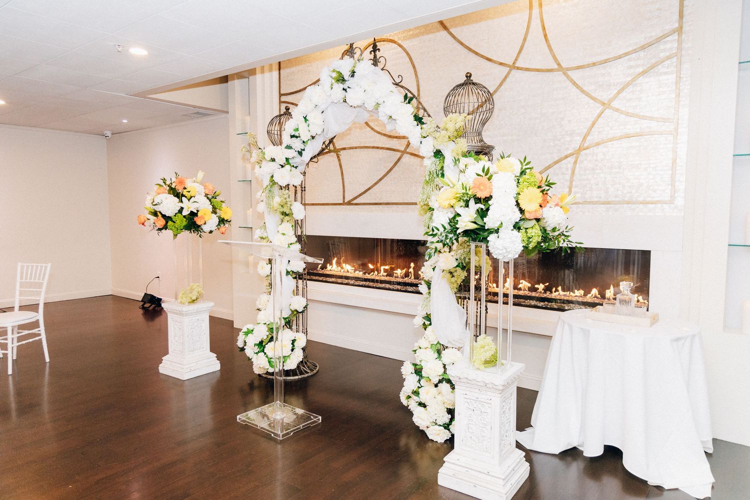 Elegant wedding ceremony setup with floral arch, white roses, and gold accents near a fireplace in a bright venue.