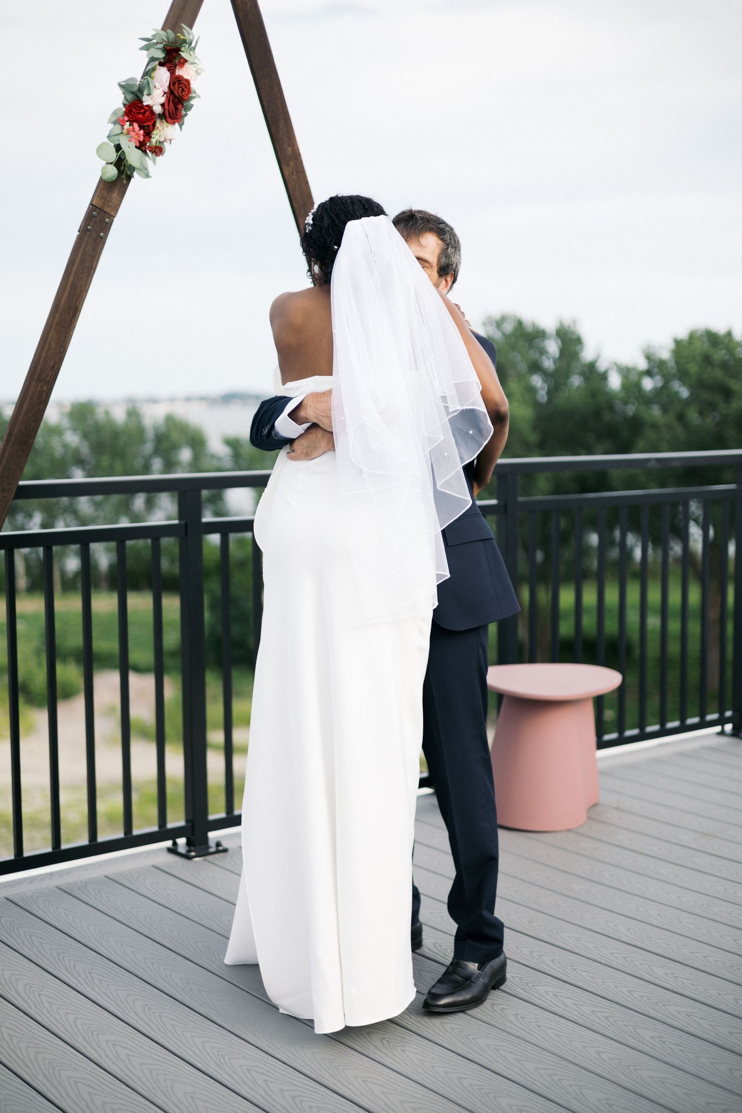 A bride and groom embrace from behind under a triangular wooden arch on an outdoor deck with scenic views.