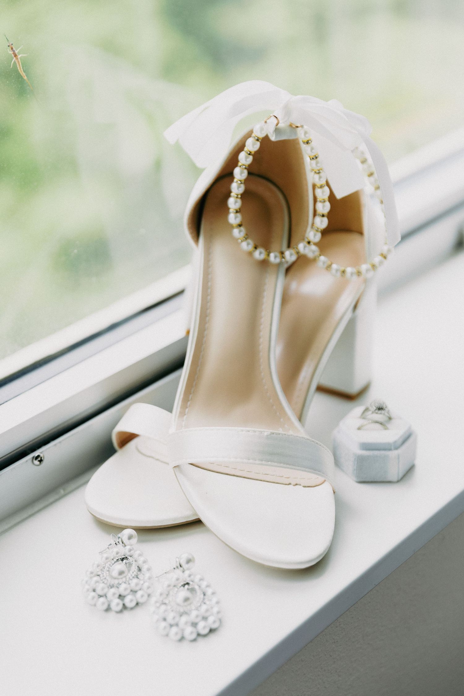 White bridal heels with pearl ankle strap and bow, displayed with crystal earrings and ring box on windowsill.