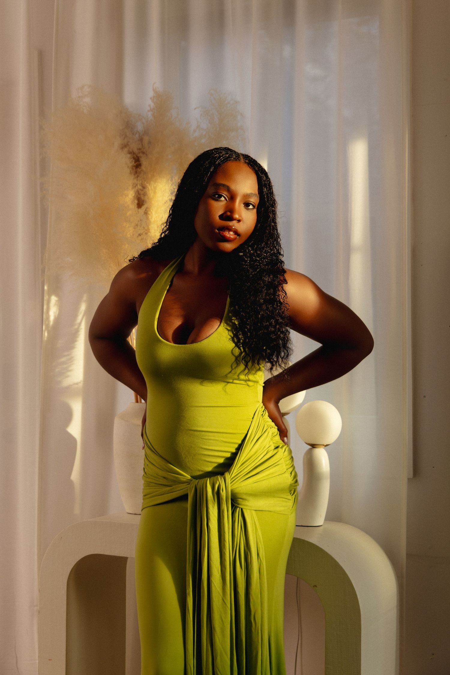 Woman in green halter maxi dress posing confidently indoors with warm sunlight and sheer curtains.