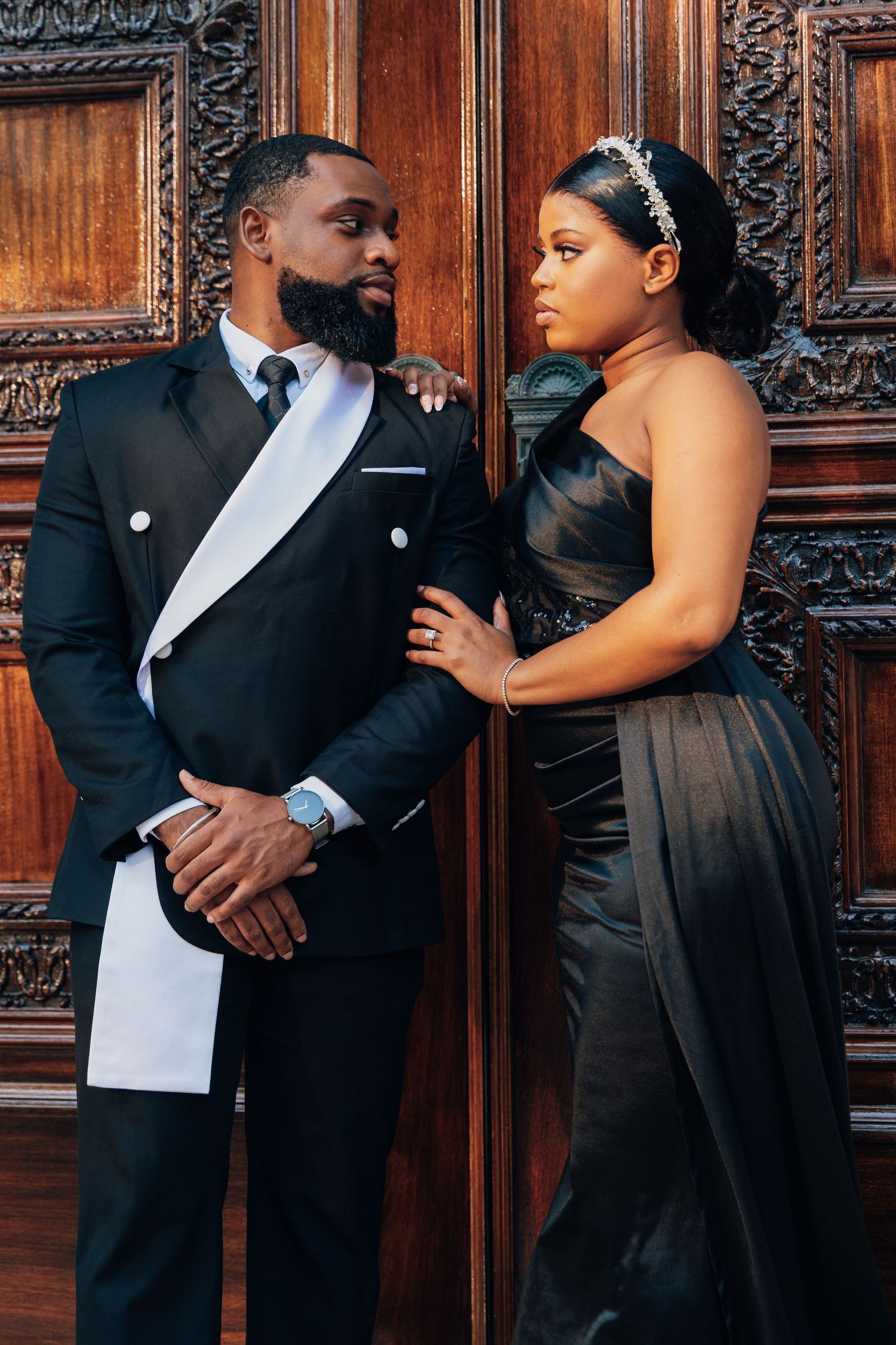 Elegant couple in formal black attire posing against ornate wooden doors, man in tuxedo and woman in strapless gown.