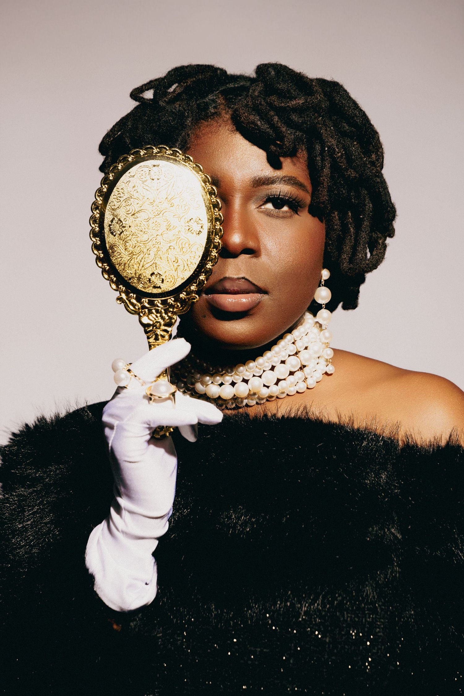 Elegant Black woman in fur coat, pearl necklace, white gloves, holding ornate gold mirror, with locs hairstyle.