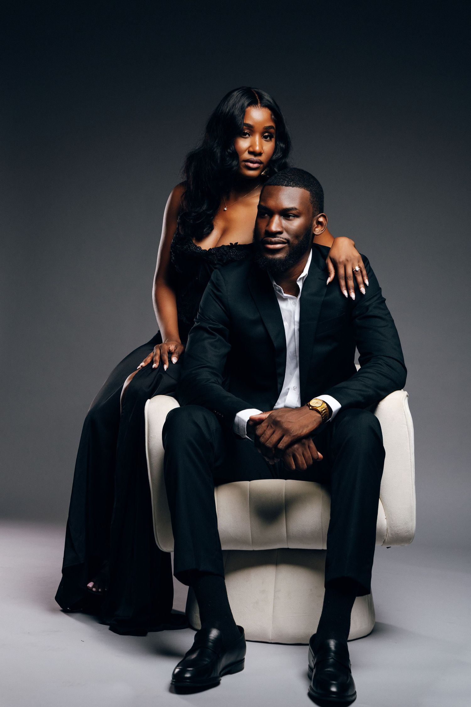 Elegant Black couple posing in studio, man seated in white chair wearing suit, woman in black gown standing behind him.